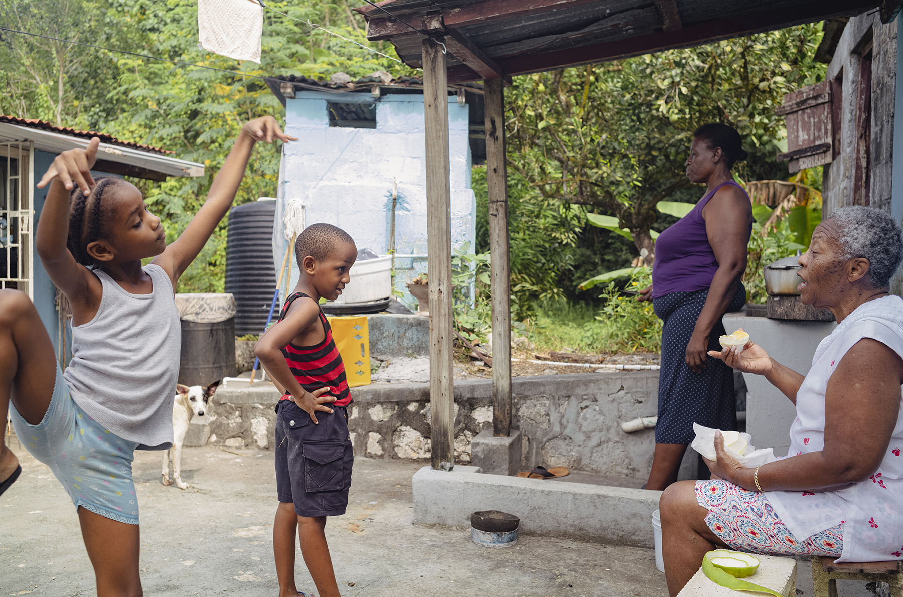 Photograph of one child dancing whilst another speaks with an older person. Another person and a dog stood in the background.