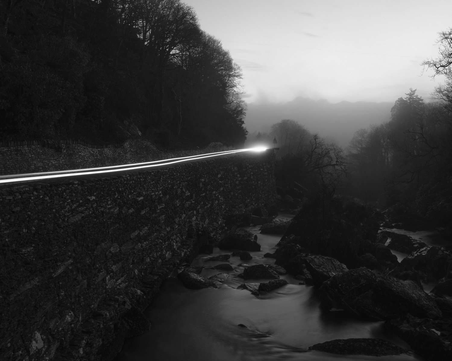 Black and white photo of a rocky stream running down alongside a road.  Car lights flash past on the road.