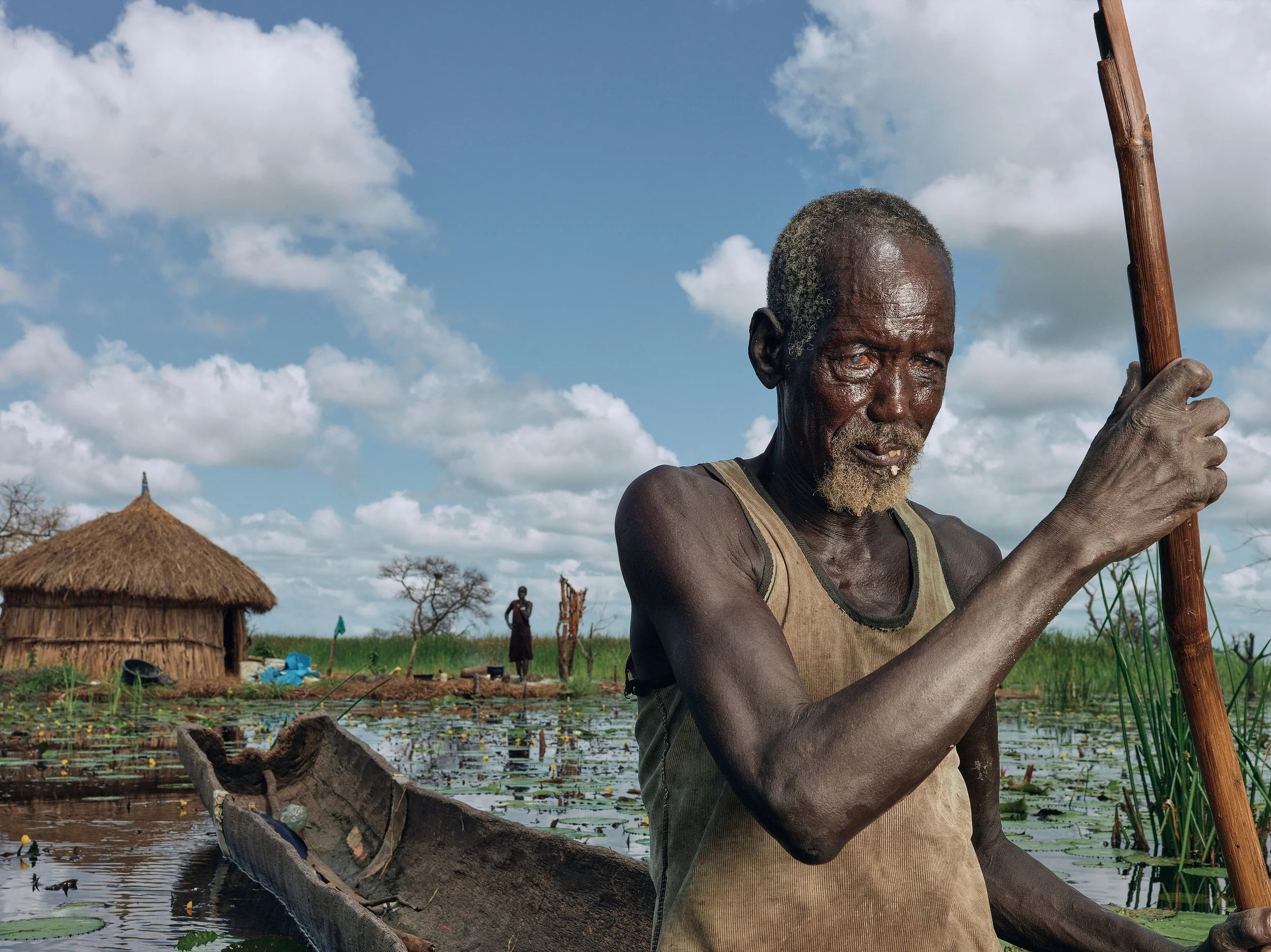 Image from exhibition of a man rowing a simple hand made canoe away from a straw hut.