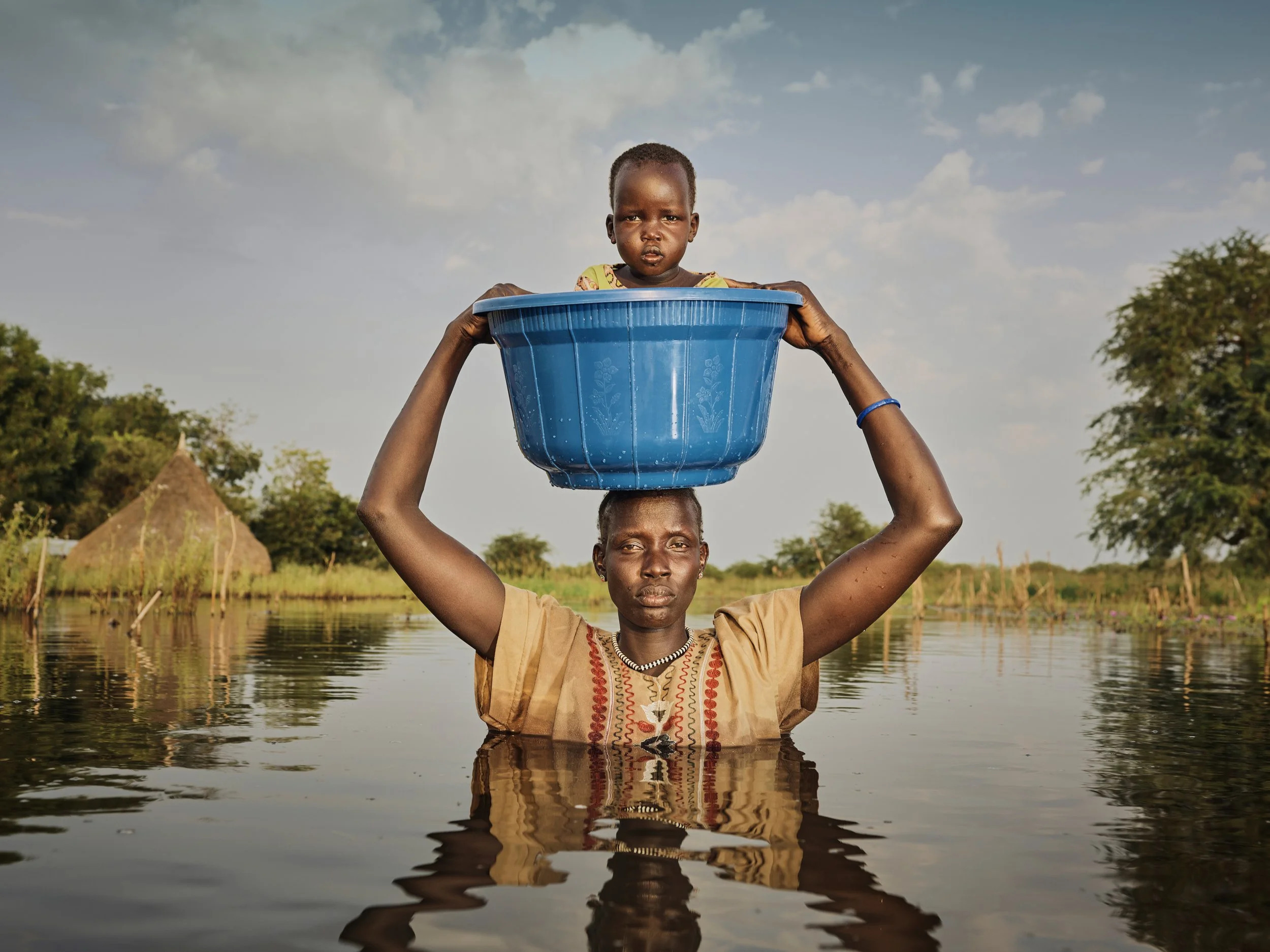 Image of a woman, chest deep in water, holding a container containing a child on her head.