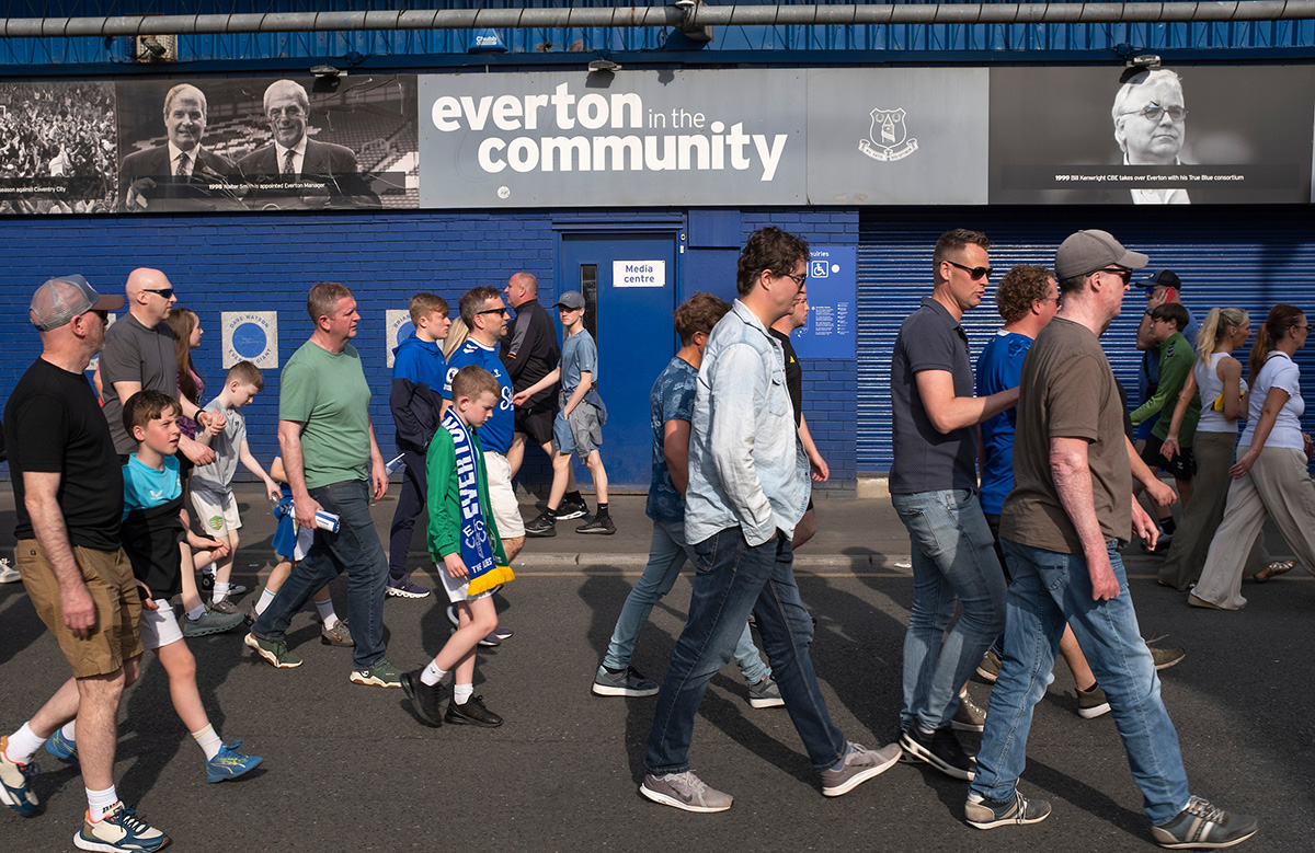 Photographer's image of a group of Everton football fans walking past a backdrop of the buildings painted in blue.  A sign above reads "Everton in the Community".