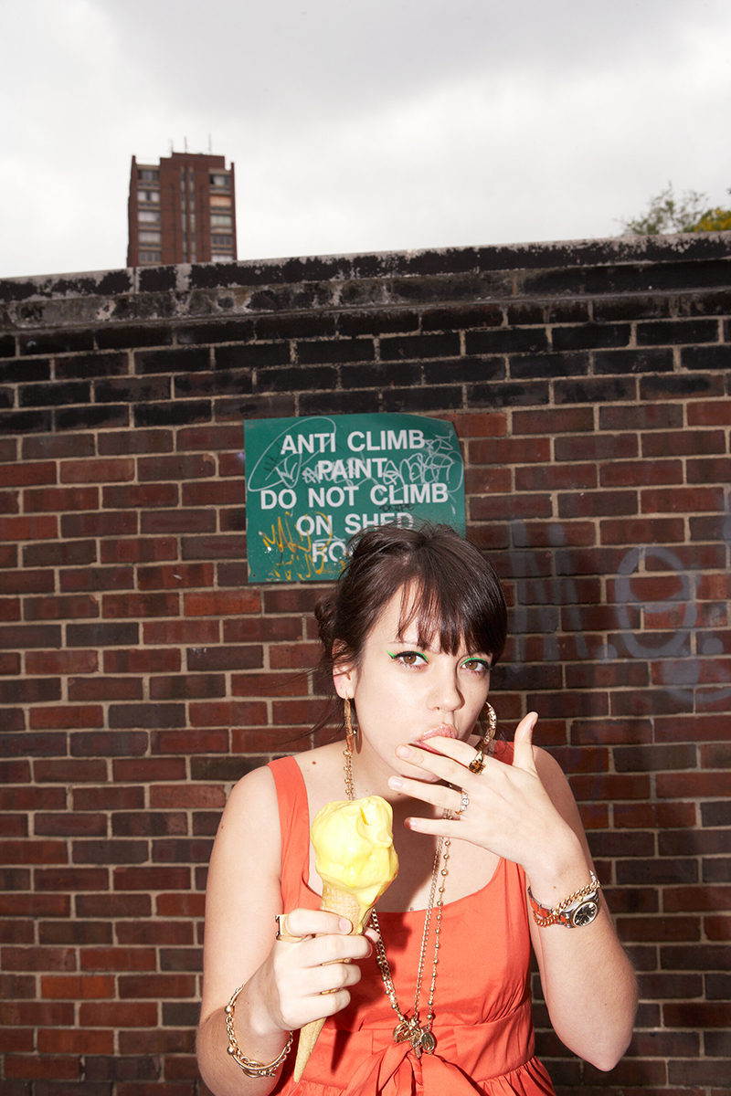 Lily Allen eating an ice cream in front of a brickwall displaying a graffiti'd sign.