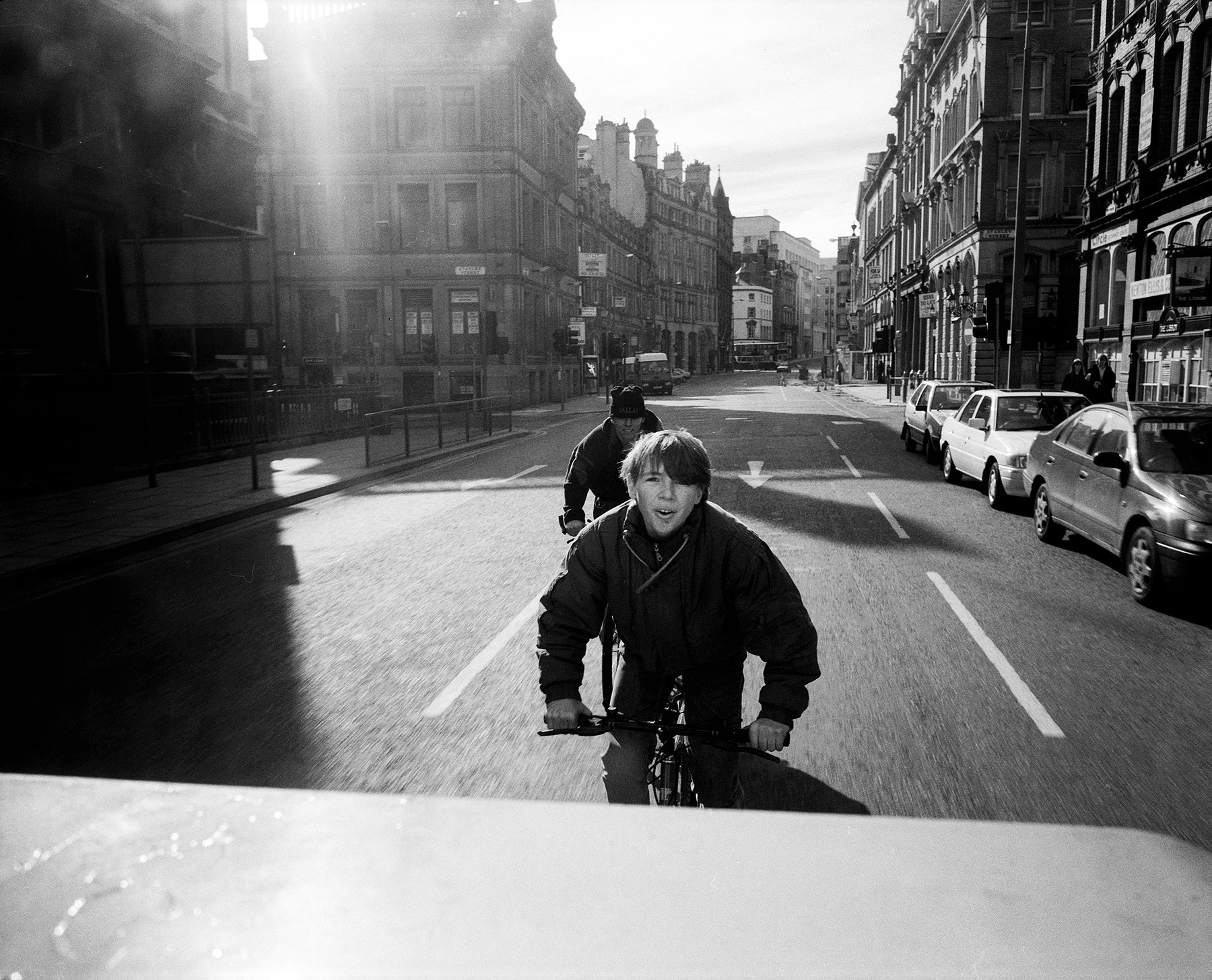 artist photograph showing a street scene with a young man cycling behind a car.