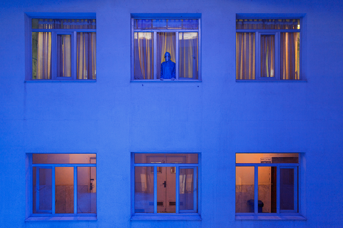 Blue lit building with 6 windows, there is a figure of a person in the top middle window